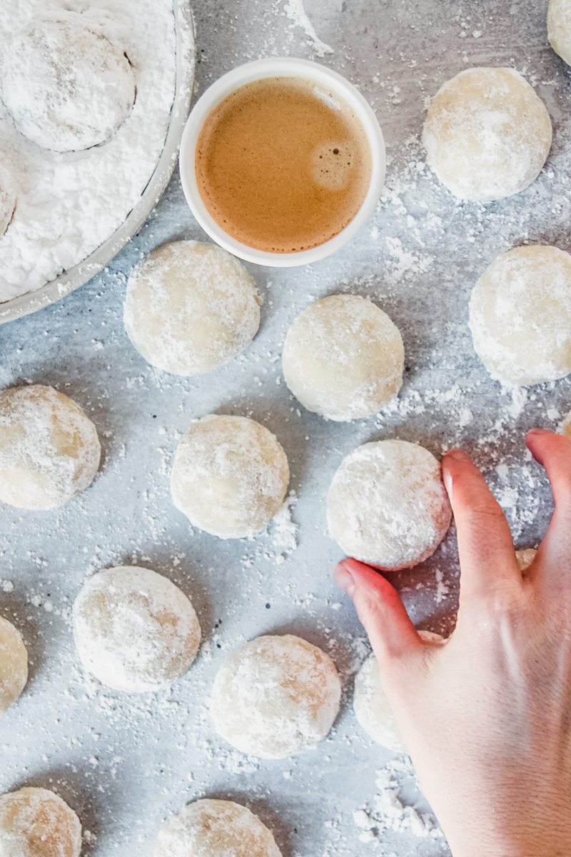 A Mexican Wedding Cookie or Polvorones is picked up from a gray surface with a cup of coffee and a rimmed ceramic plate with powdered sugar beside.