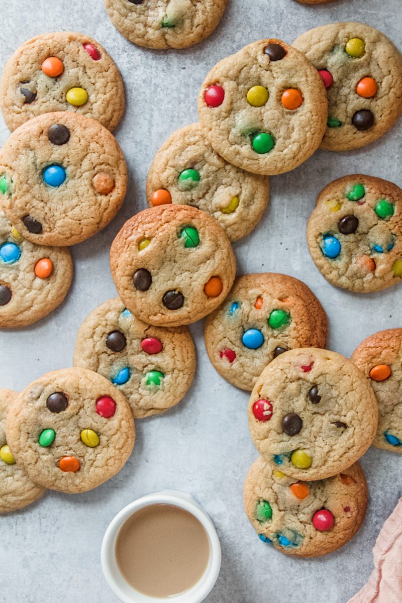 M&M cookies lay unevenly on a gray surface with a small cup of tea beside.