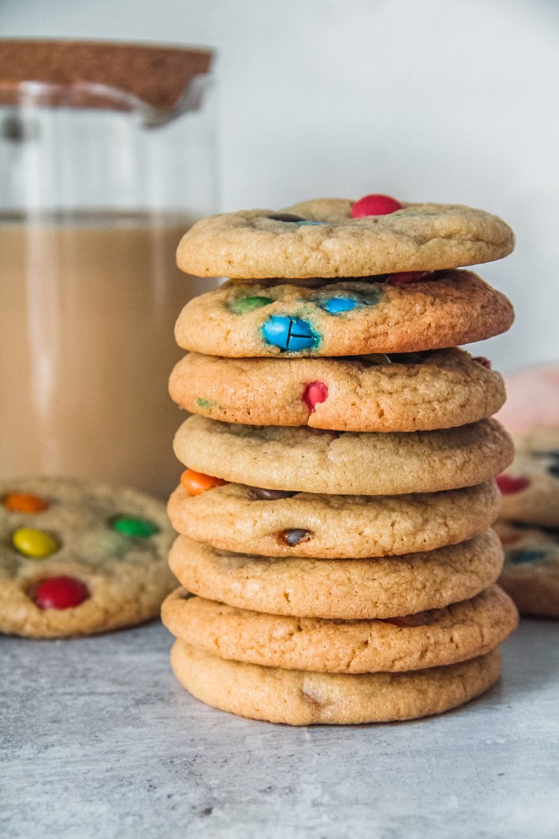 A stack of M&M cookies sit on a gray surface with a glass jug of tea in the background.