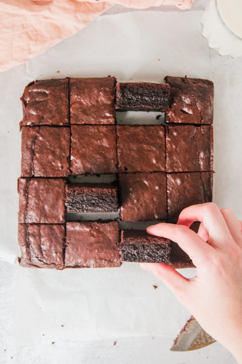 A piece of brownie is pucked up from a gray surface surrounded by a tray of brownies laid out around it.