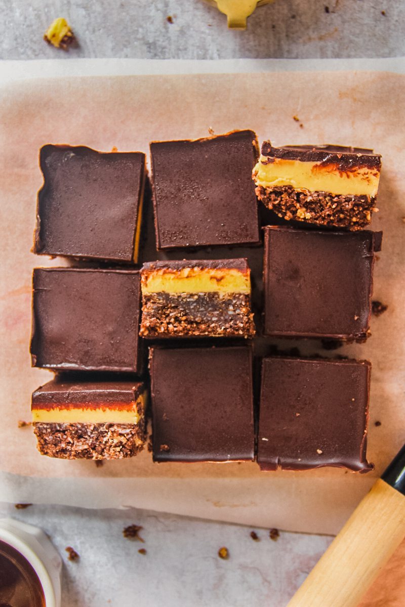 Nanaimo bars sit on a lined wooden board on a gray surface with some turned sideways to show the three layers with a small cup of coffee beside.