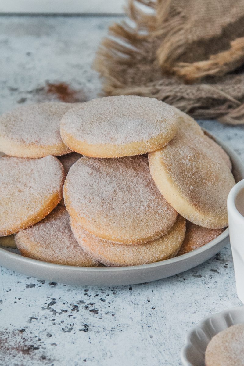 Sugar coated Biscochito cookies sit stacked on a rimmed ceramic gray plate on a gray surface.