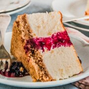 A slice of New York Cheesecake sits on an individual plate on its side topped with a berry sauce on a gray surface.