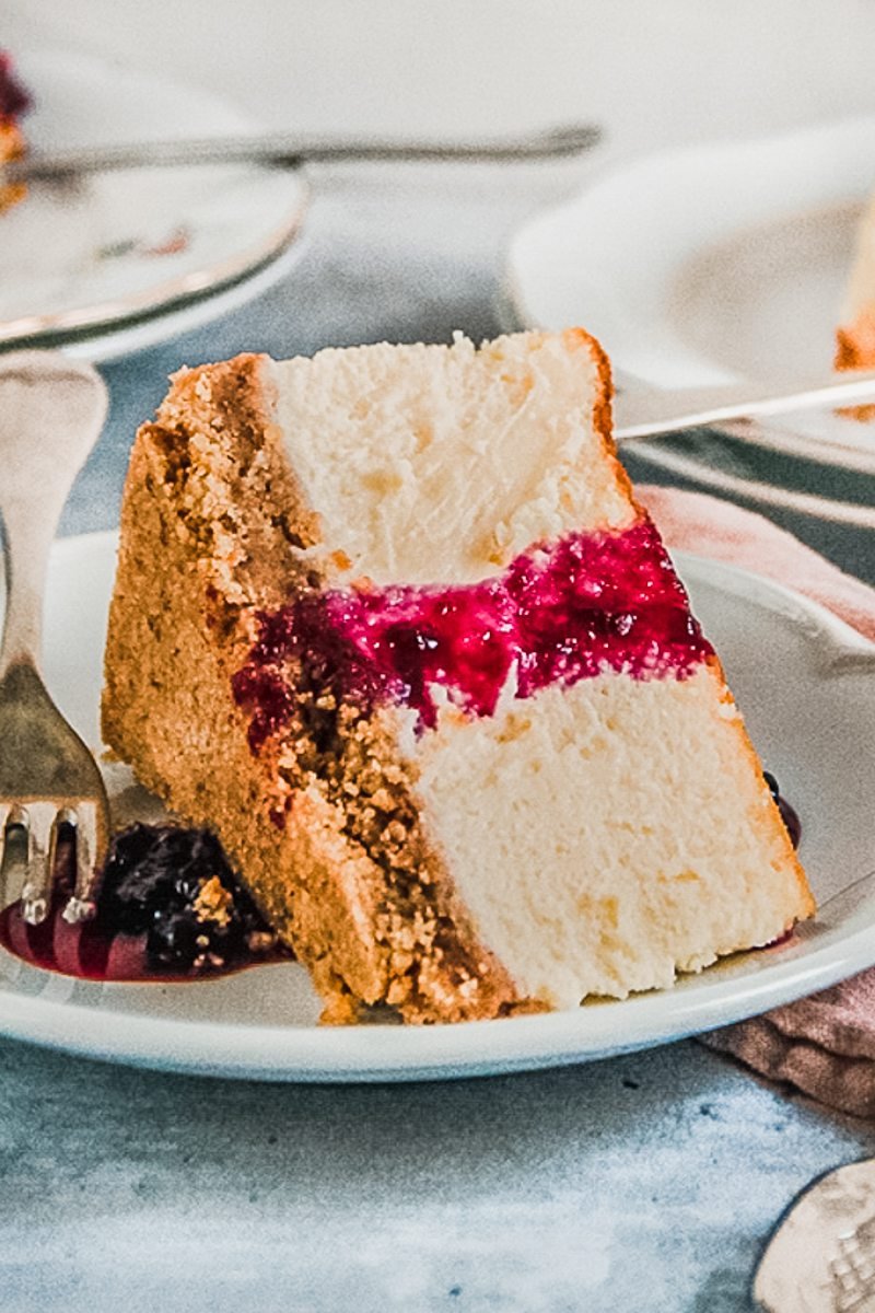 A slice of New York Cheesecake sits on an individual plate on its side topped with a berry sauce on a gray surface.