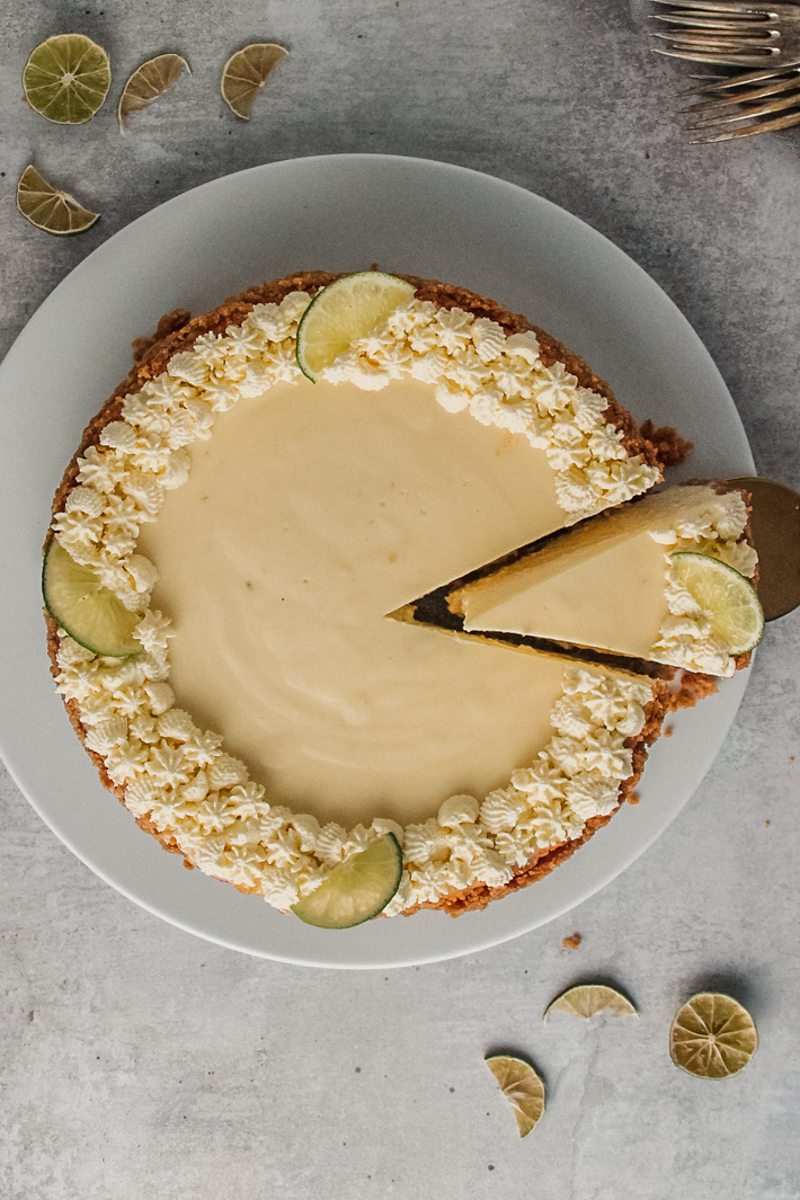 A slice of Key lime cheesecake sits on a white ceramic plate beside the remaining cake.