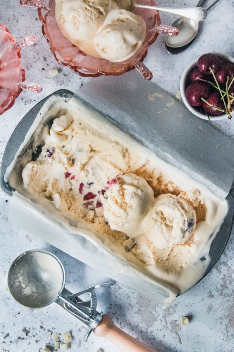 Scoops of no churn white chocolate cherry semifreddo sits in a metal loaf tin on a light grey surface with a pink glass bowl filled with melting scoops and cherries above.