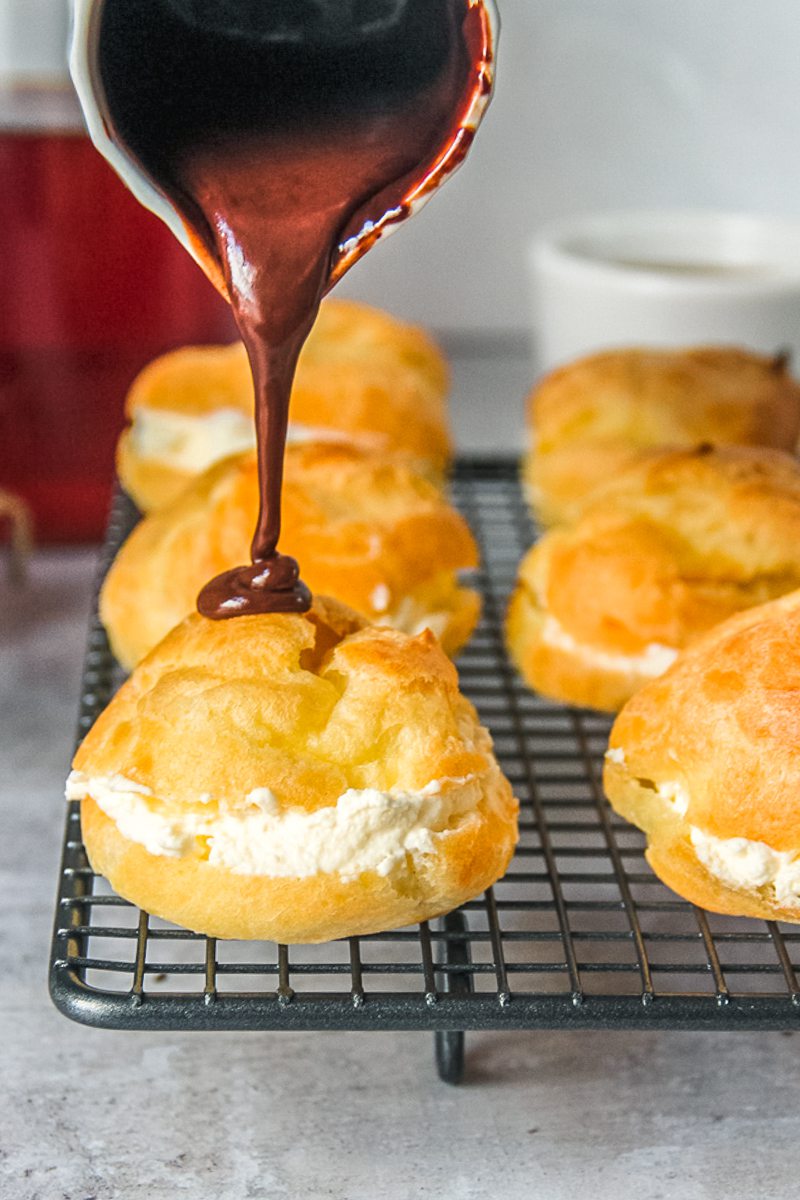 Chocolate sauce is poured over a cream filled choux pastry on a cooling rack.