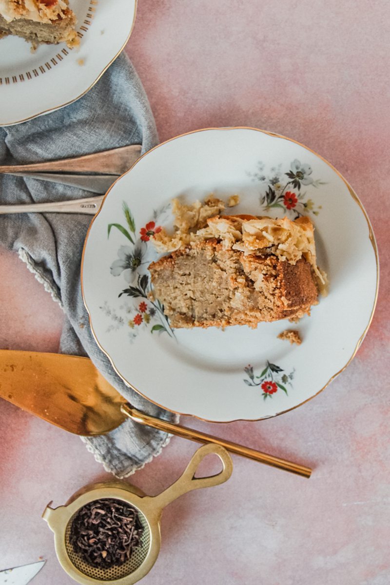 A slice of oatmeal cake with a coconut topping sits on a white ceramic plate on a light pink surface.