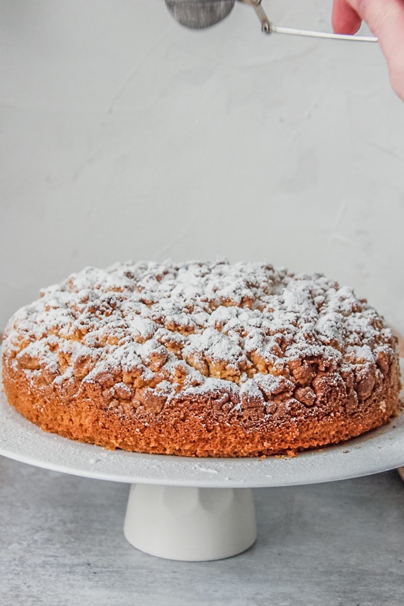 Powdered sugar is sprinkled over a dairy free olive oil cake sitting on a cake stand on a gray surface.