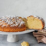A slice of Olive Oil cake sits in a serving knife from a cake stand on a gray surface.