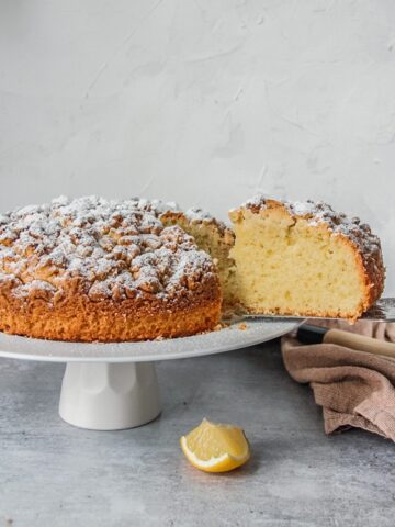 A slice of Olive Oil cake sits in a serving knife from a cake stand on a gray surface.