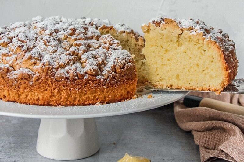 A slice of Olive Oil cake sits in a serving knife from a cake stand on a gray surface.