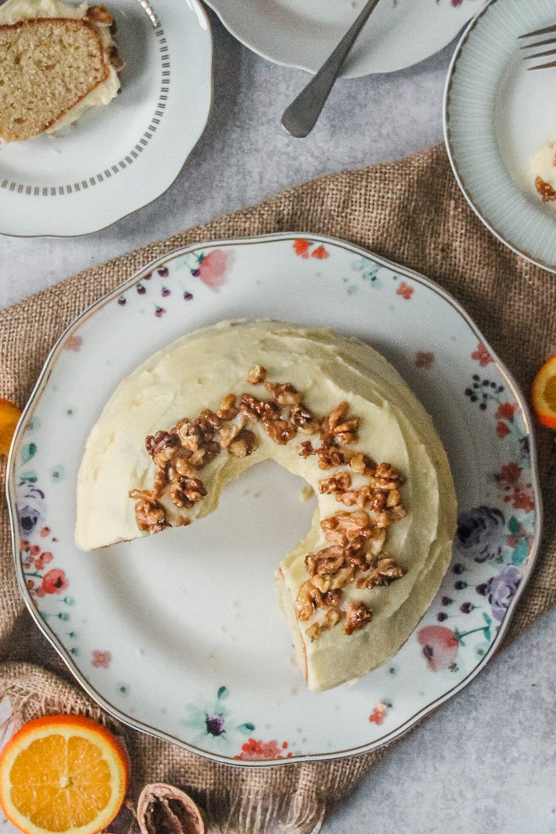 An orange bundt cake sits on a white floral ceramic plate on a gray surface on a brown cloth with slices sitting on individual plates beside.