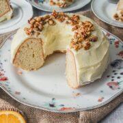 An orange bundt cake with orange frosting and maple walnuts sits on a white floral ceramic plate on a gray surface on a brown cloth with slices sitting on individual plates beside.