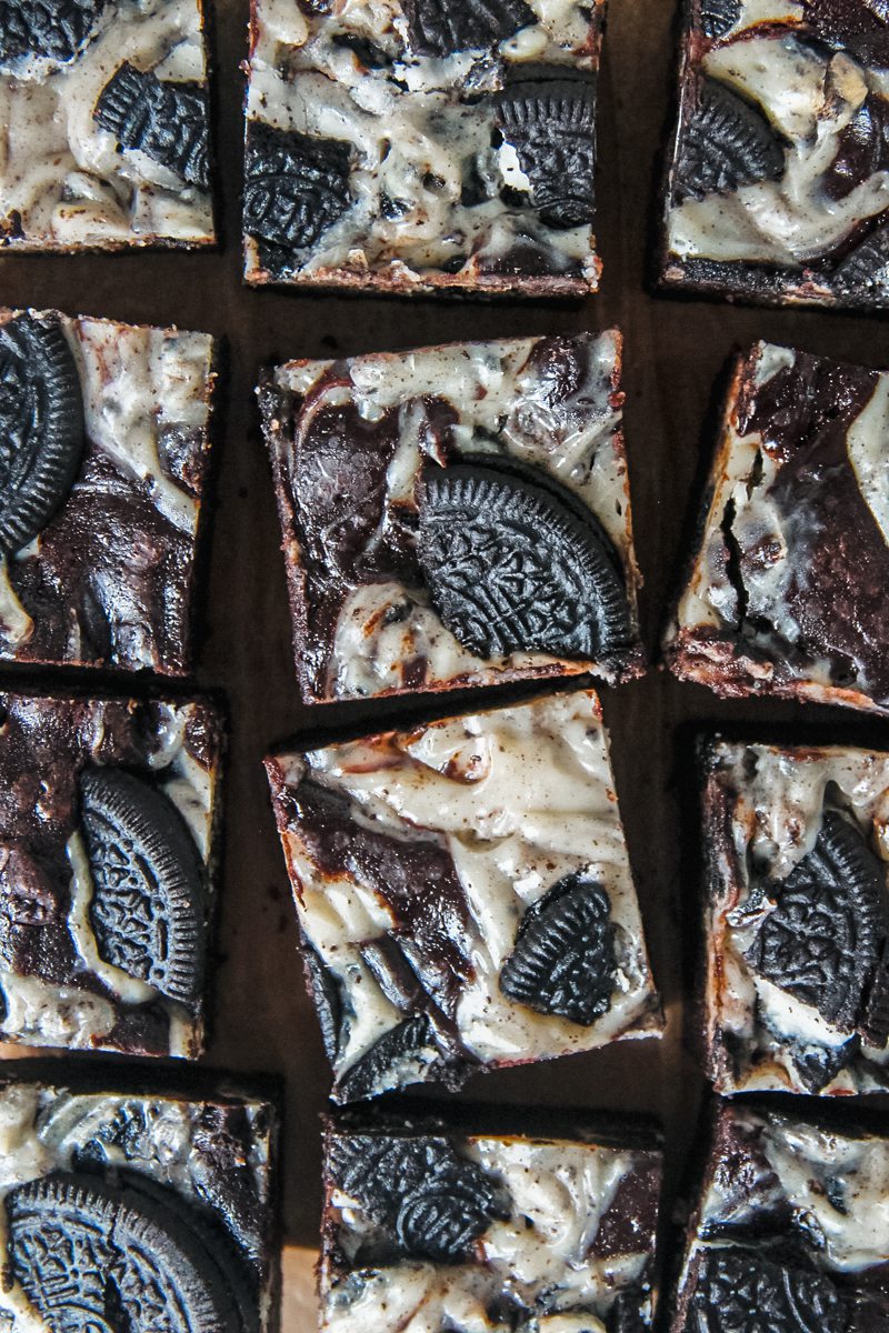 Squares of oreo cheesecake brownies with pieces of oreos on a lined surface.