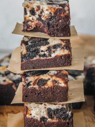 A stack of Oreo cheesecake brownies sit on each other on a parchment paper surface with the top brownie having had a bite taken out of it.