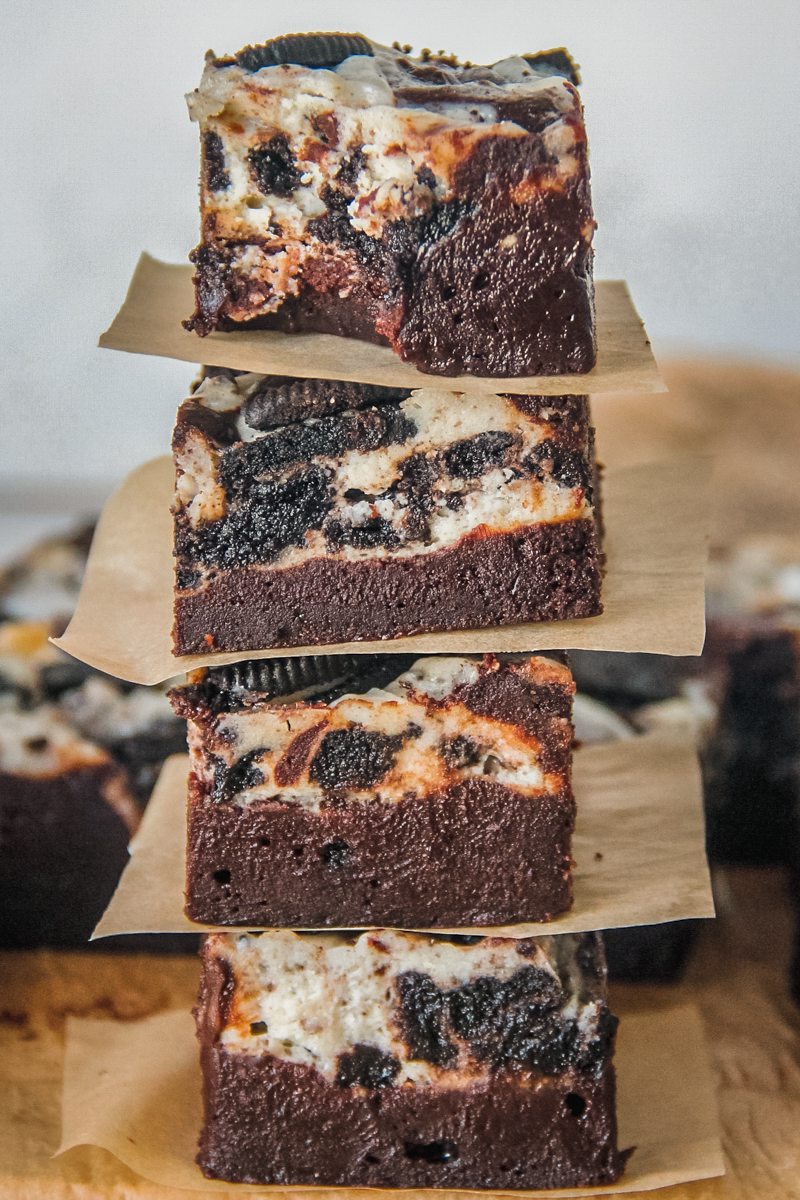 A stack of Oreo cheesecake brownies sit on each other on a parchment paper surface with the top brownie having had a bite taken out of it.