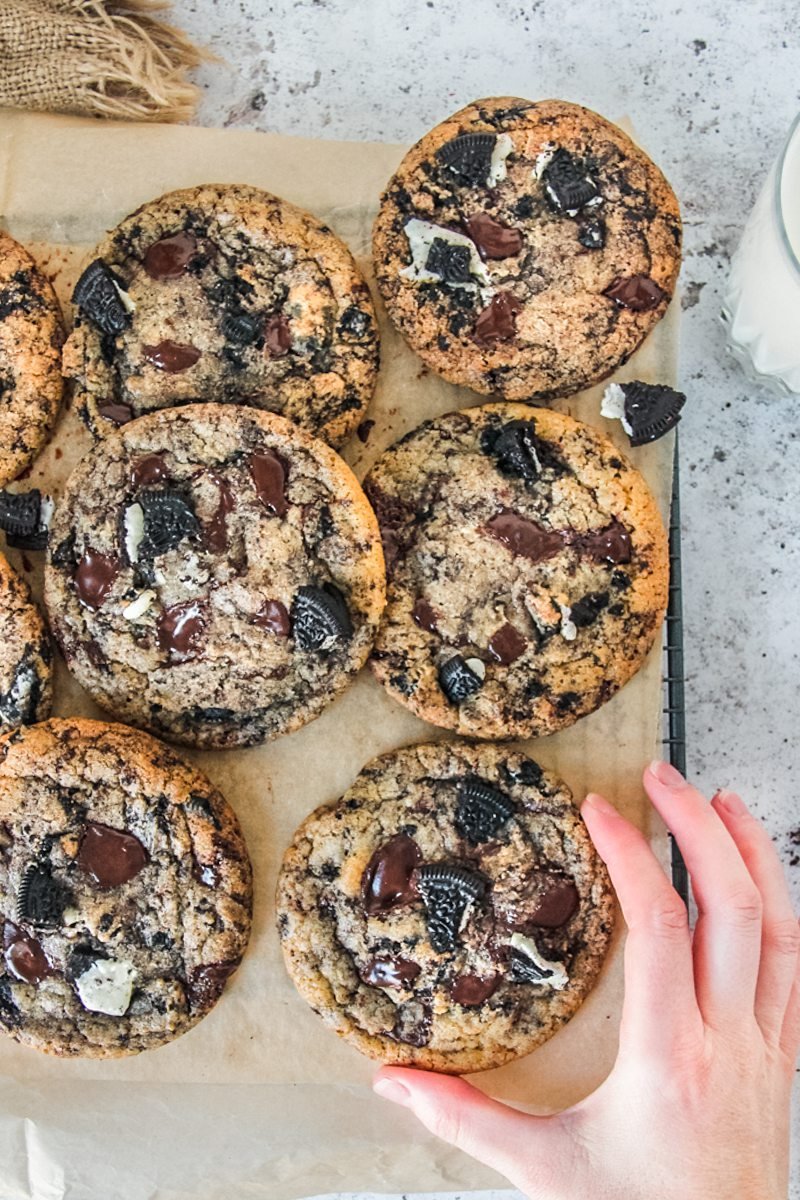 An oreo cookies and cream cookie is about to be picked up from a cooling rack lined with parchment paper on a light gray surface.