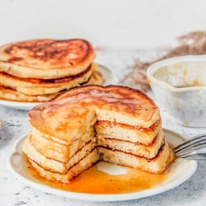 A stack of lemon pancakes sits cut with a wedge on a white ceramic plate on a light gray surface.