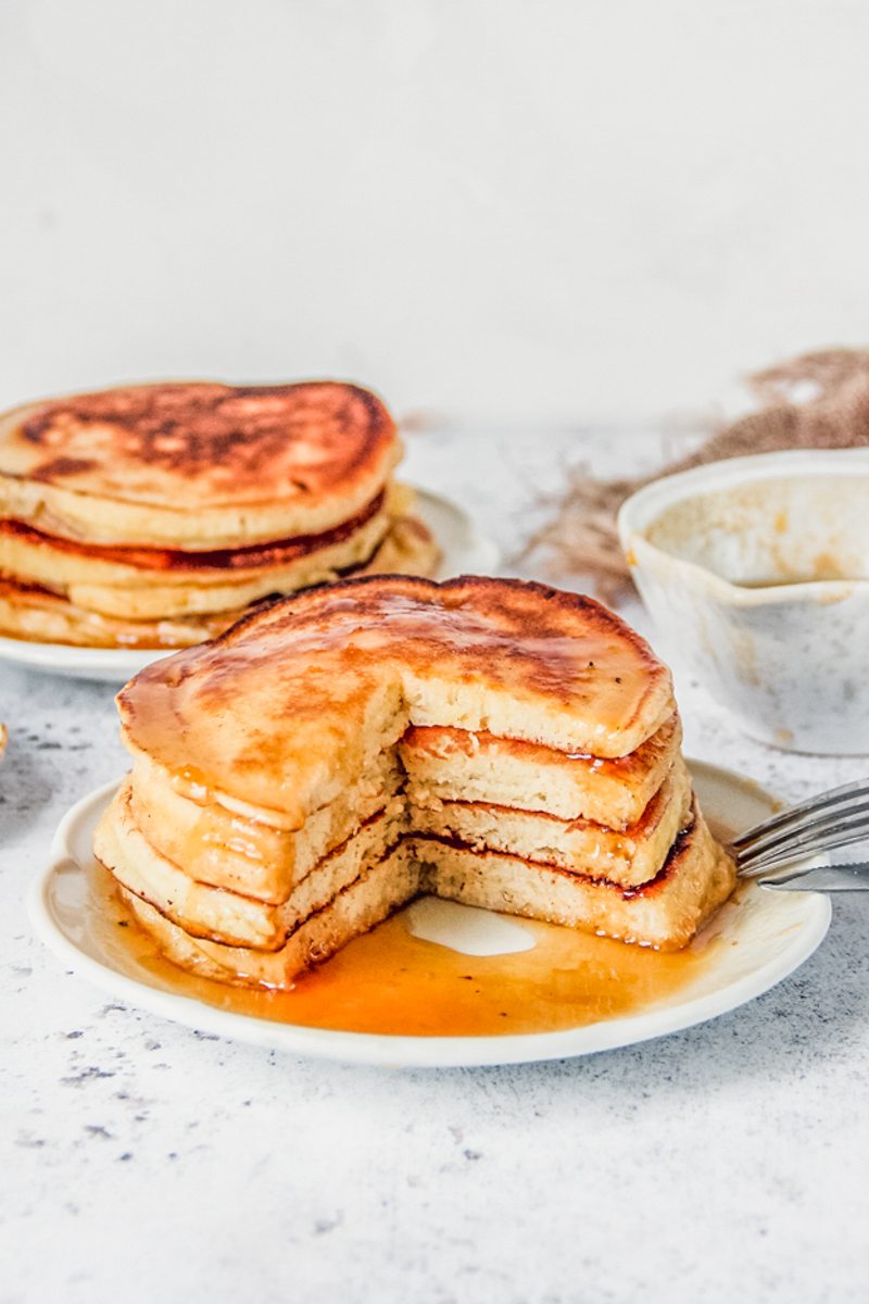 A stack of lemon pancakes sits cut with a wedge on a white ceramic plate on a light gray surface.