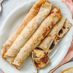 A spoonful of rolled pancakes filled with custard sits on a plate of rolled custard filled pancakes on an oval white ceramic plate on a gray surface.