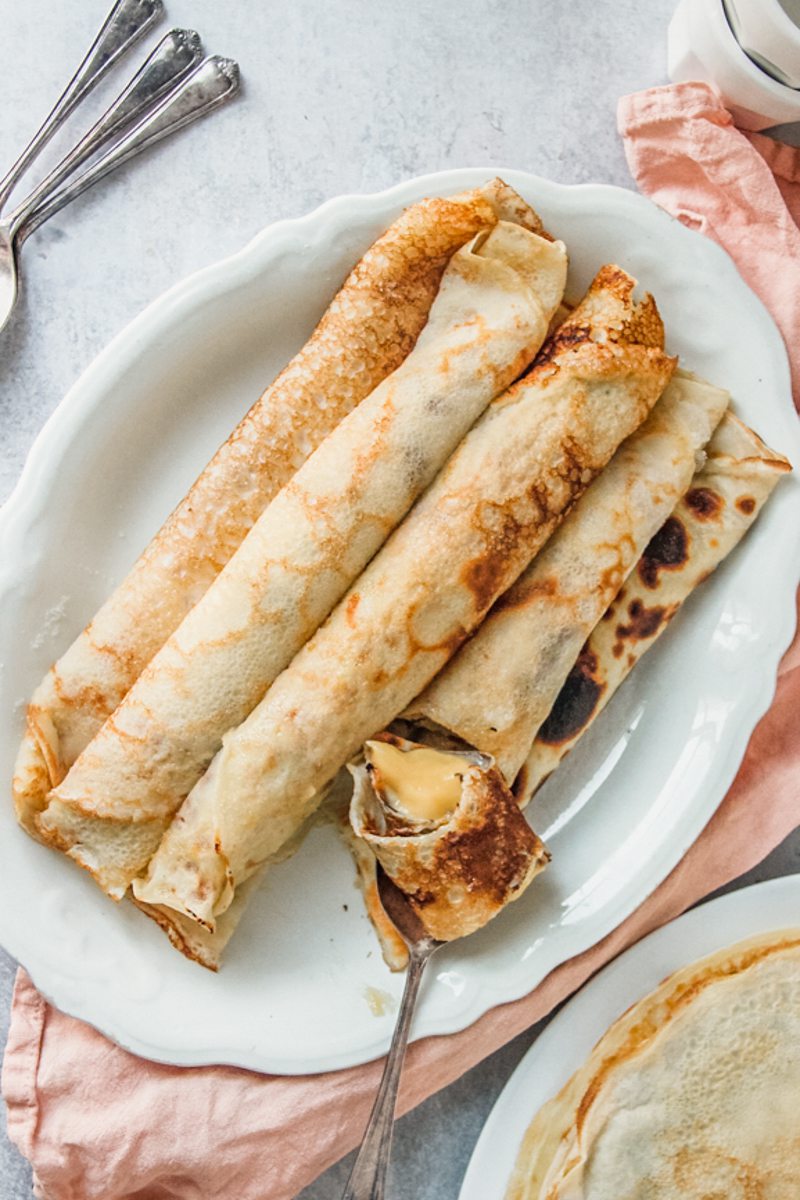 A spoonful of rolled pancakes filled with custard sits on a plate of rolled custard filled pancakes on an oval white ceramic plate on a gray surface.