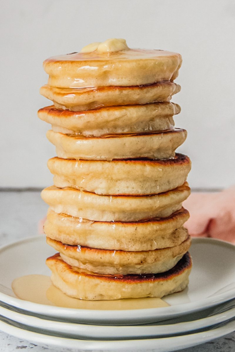 A stack of griddle cakes sit on a stack of white ceramic plates with melting butter and drizzled honey.