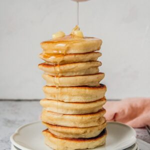 Honey is poured over a stack of Scotch cakes sitting on a stack of white ceramic plates on a light gray surface.