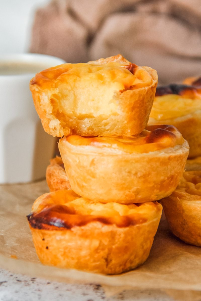 Pasteis de Natas or Portuguese Tarts sit stacked on parchment paper on a light gray surface.