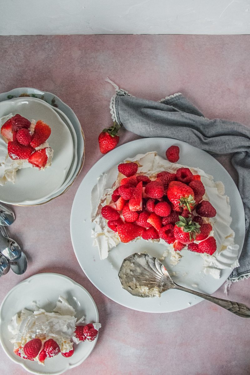 A pavlova sits on a white ceramic plate topped with strawberries & rasberries on a light pink background with two servings alongside on individual plates.