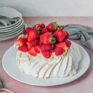 A pavlova sits on a white ceramic plate topped with strawberries & rasberries on a light pink background.