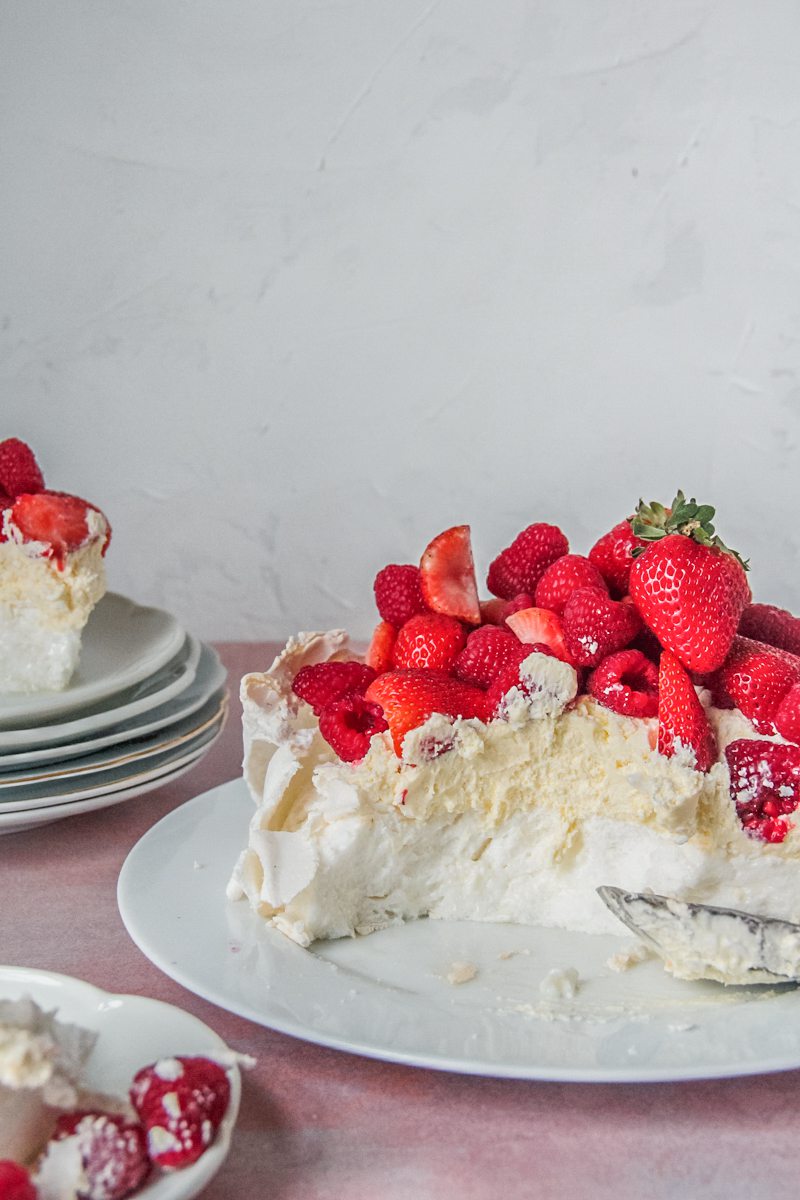 The marshmallow interior of a pavlova sits on a white ceramic plate topped with a lemon curd cream, fresh strawberries & rasberries on a light pink background.