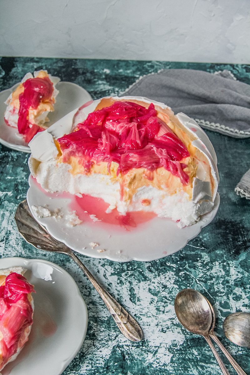 A side image of marshmallow centered pavlova topped with a mascarpone custard and rhubarb on a white plate on a green and white speckled surface.