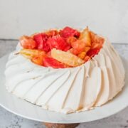A pavlova is topped with roasted segments of winter citrus on a white ceramic plate on a wooden cake stand on a light gray surface.