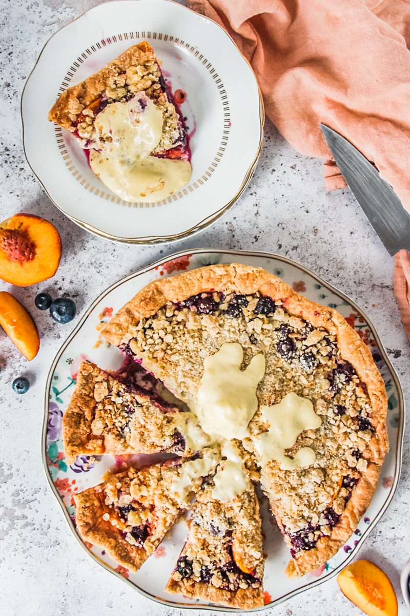 A peach and blueberry pie with streusel topping sits on a floral ceramic plate with slices cut out and a slice beside on an individual plate on a light gray surface.