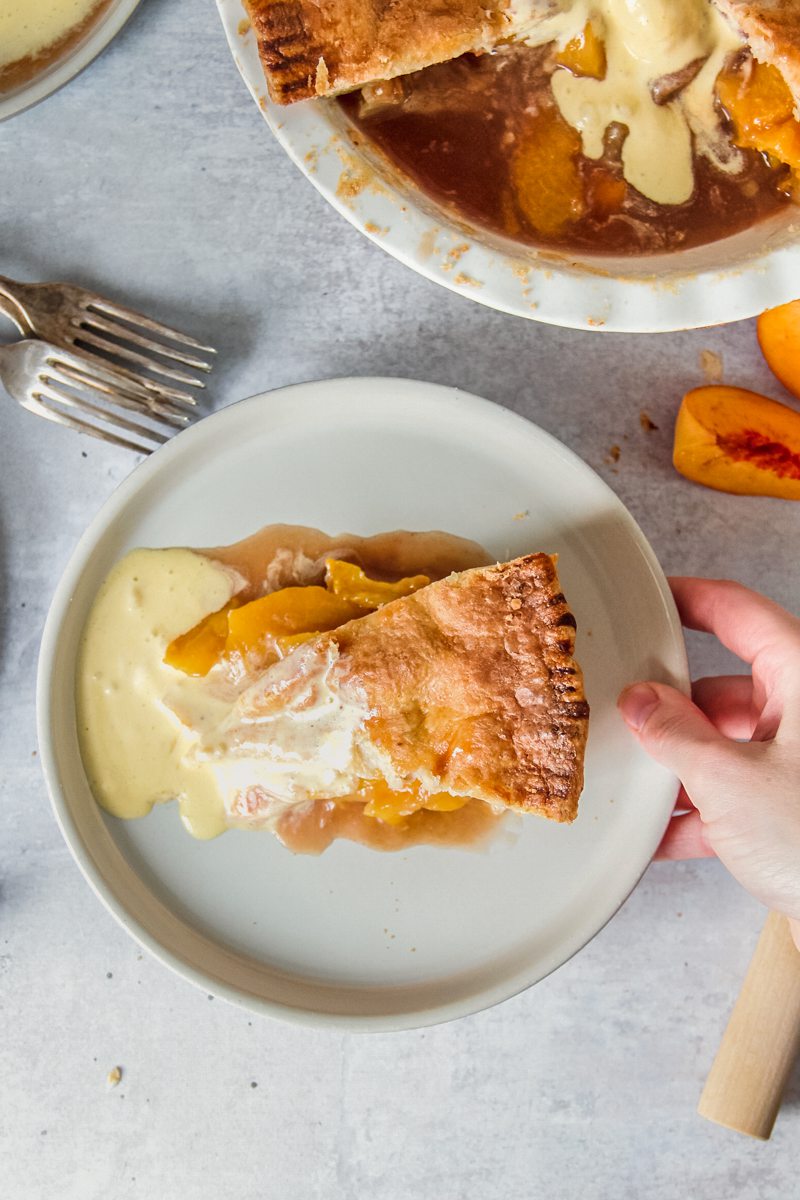 An individual serving of double crusted peach cobbler is held above the remaining pie with melty vanilla ice cream mixed with the peach juices above a gray surface.