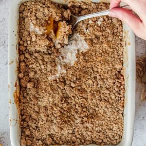 Peach crumble or peach crisp has a serving spoon lifting up a portion of the dessert from a white rectangle baking dish on a gray surface.