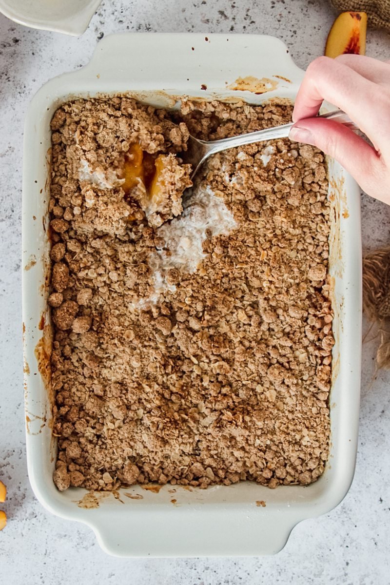 Peach crumble or peach crisp has a serving spoon lifting up a portion of the dessert from a white rectangle baking dish on a gray surface.