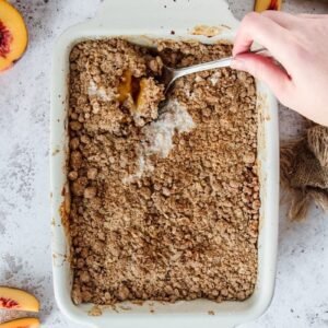 Peach Crumble is dug into with a serving spoon while it sits on a light gray background.