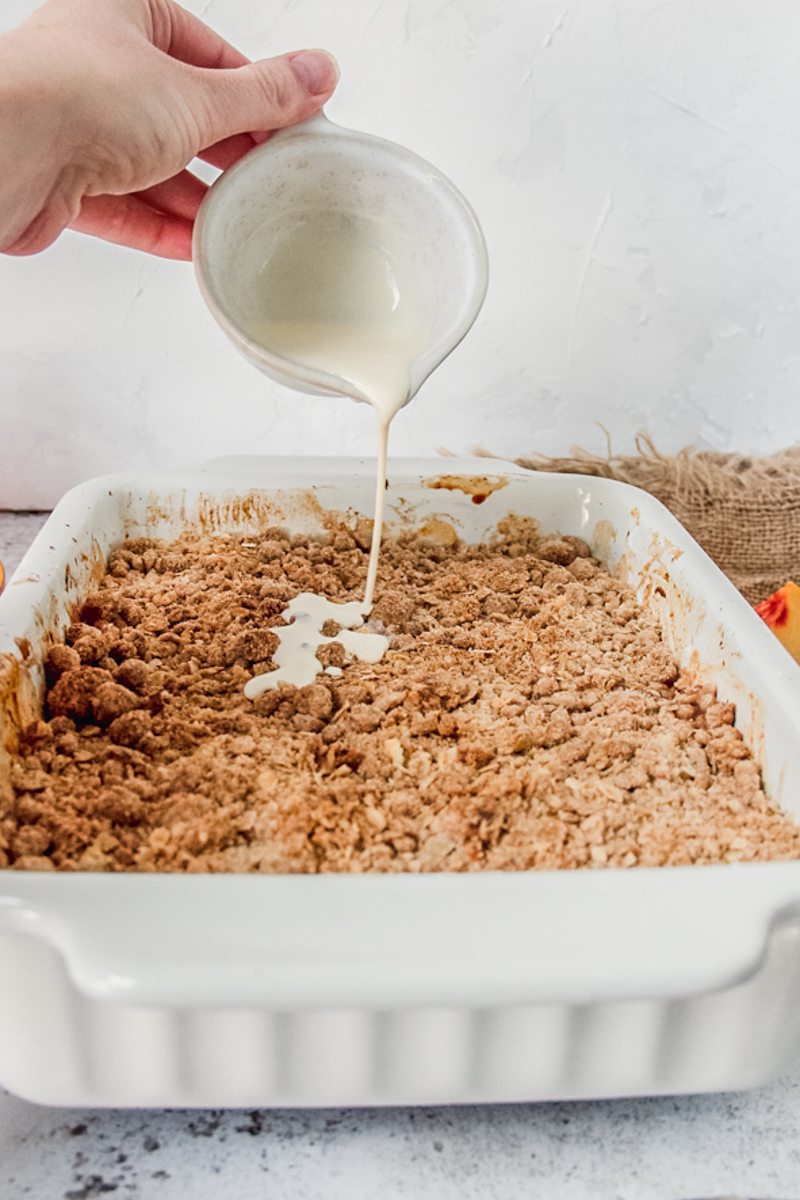 Peach crumble or crisp sits in a white rectangle baking dish on a gray surface with pouring cream being drizzled on top.