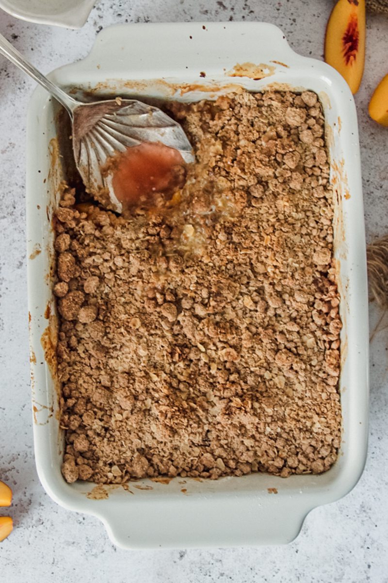 A serving spoon sits in peach crumble or peach crisp from a white rectangle baking dish on a gray surface.