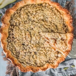 A slice of pear streusel pie sits on top of two cloths on a blue gray surface with halved pears beside with a slice being lifted on a cake server.