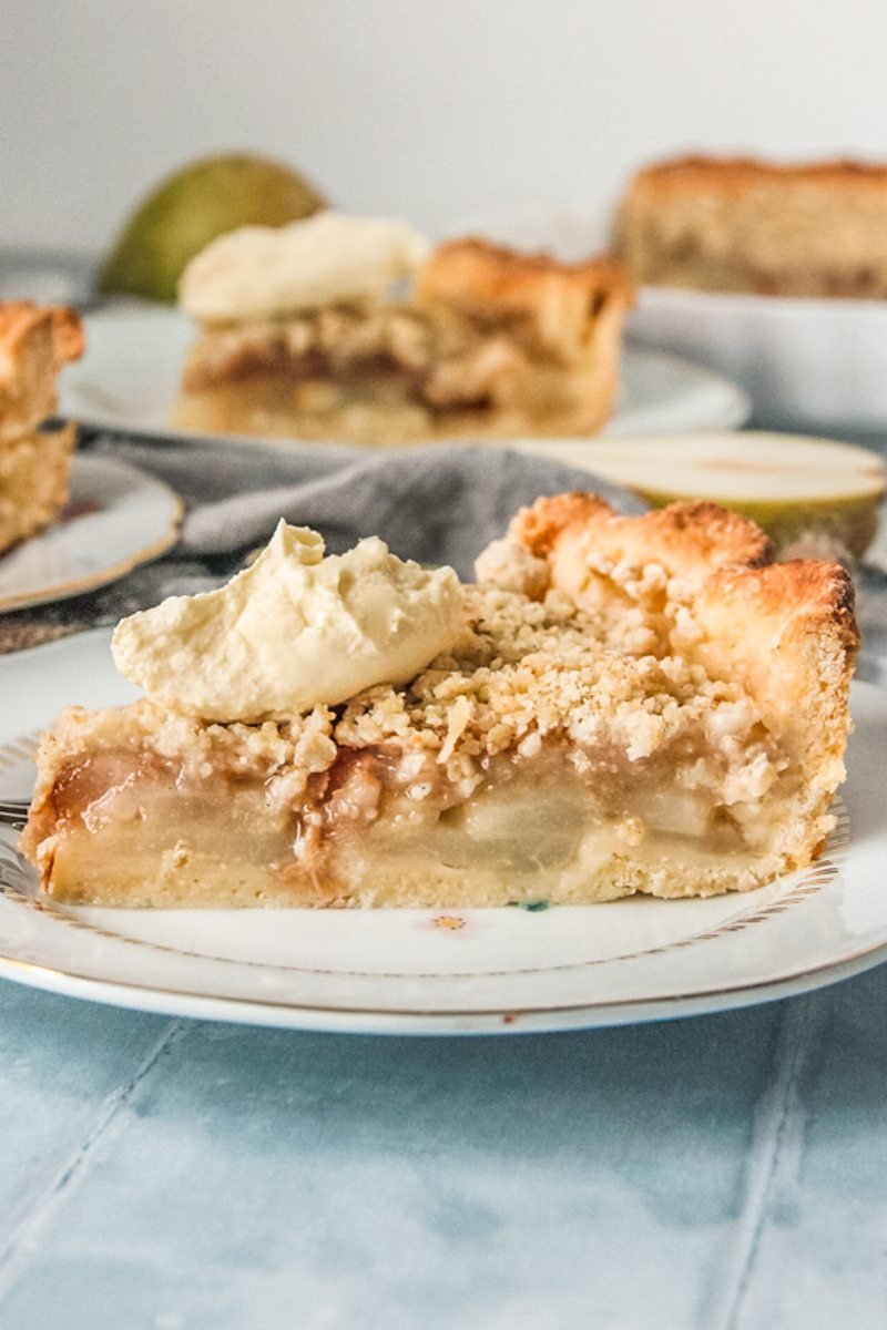 A slice of pear crumble pie sits on an individual serving ceramic plate with a dollop of whipped cream on top.