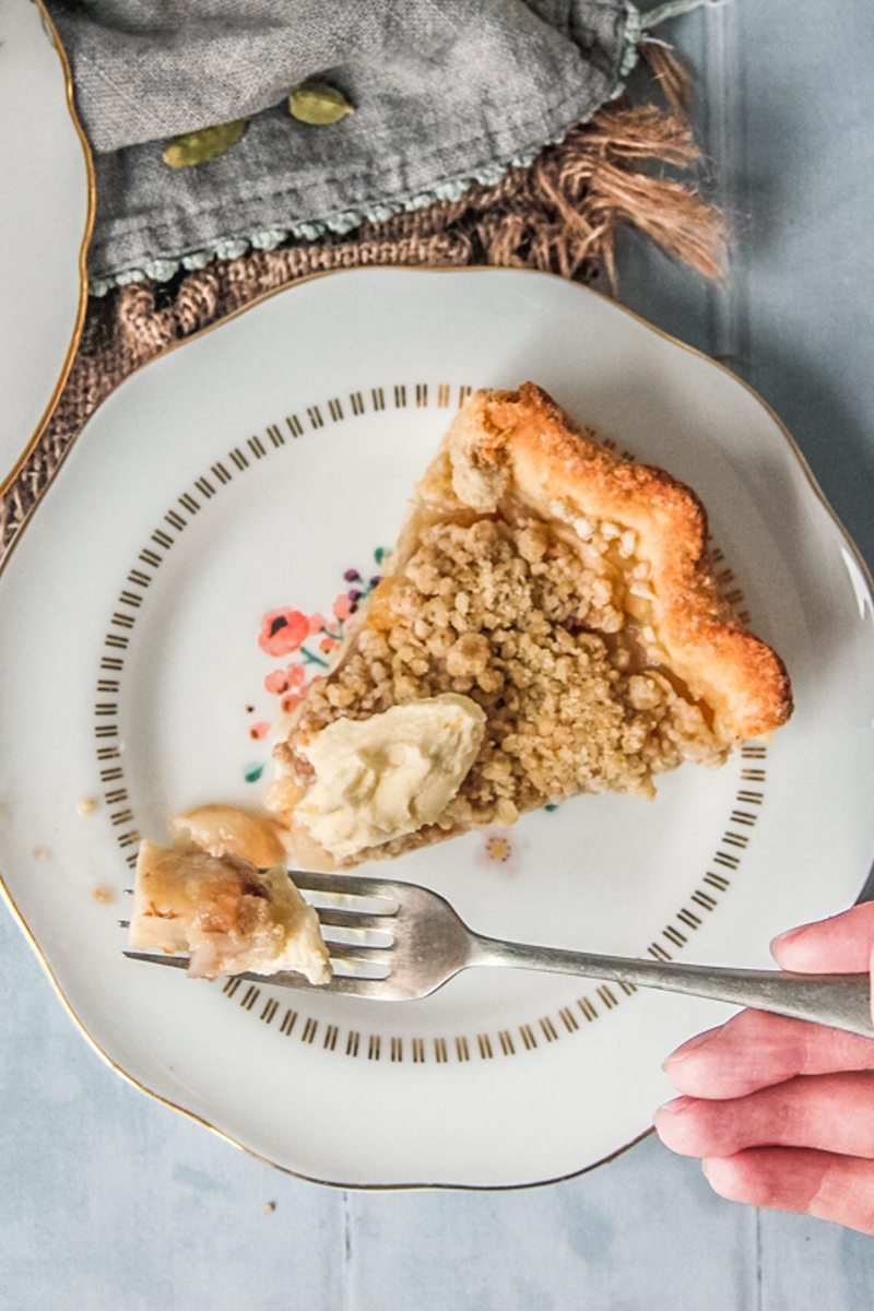 A slice of pear pie sits on an individual ceramic plate on a blue gray surface with a forkful being served up.