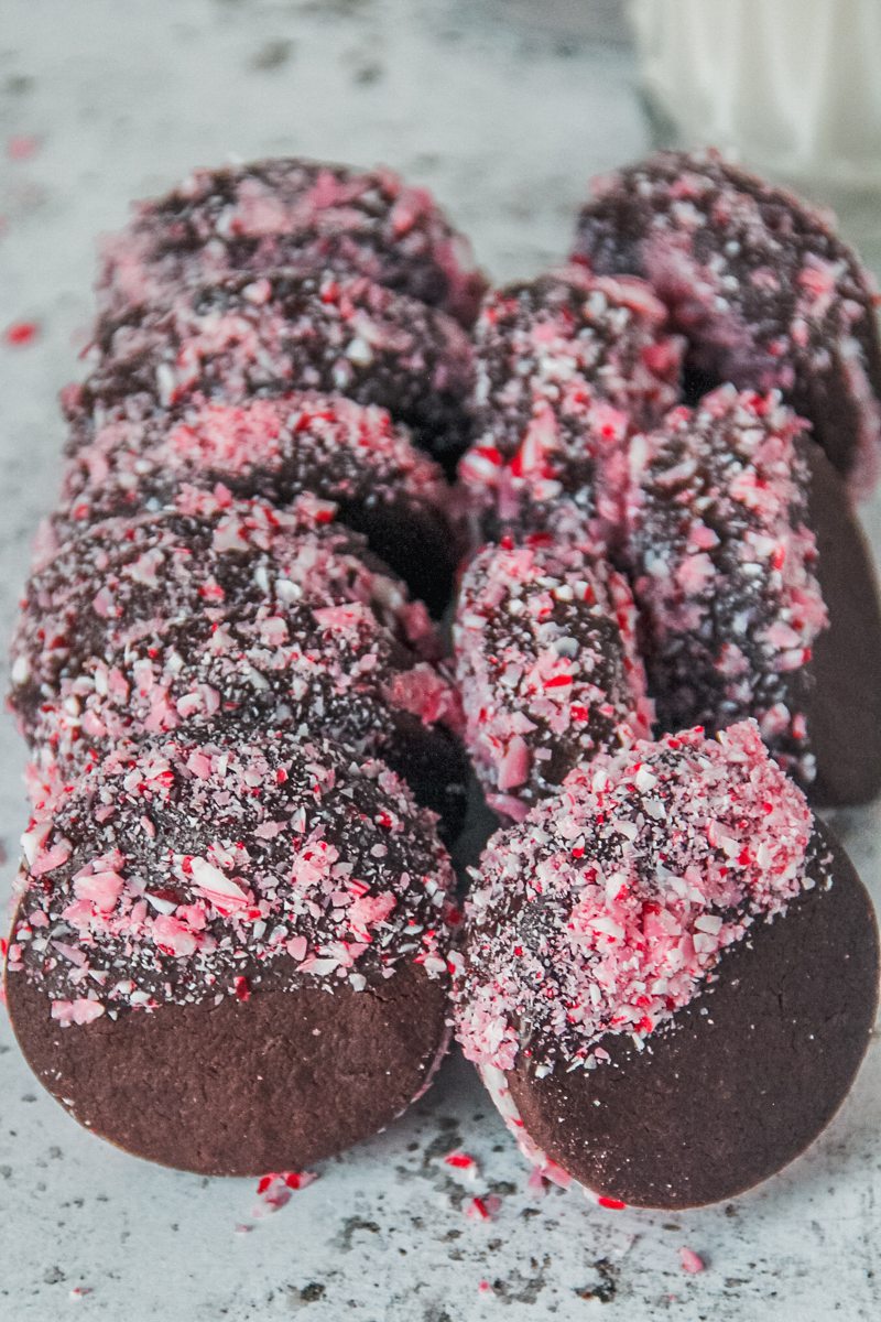 Chocolate peppermint sandwich cookies decorated with crushed candy canes rest on other cookies on a light gray surface.