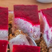 Squares of cranberry cheesecake with a crushed biscuit base, no bake filling and layer of cranberry puree sit on parchment paper with a spoonful of the cheesecake resting on a spoon beside.