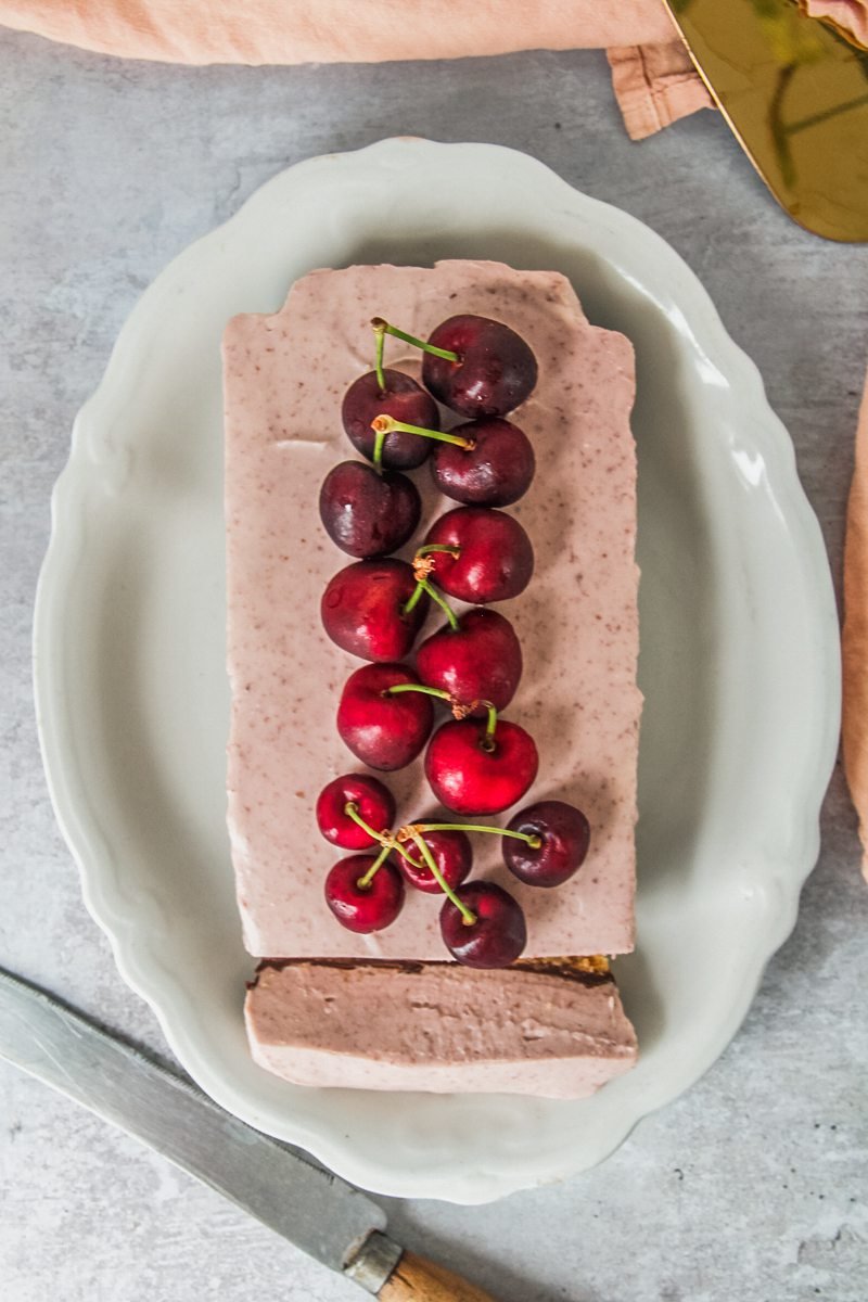 A cherry cheesecake sits on an oval white ceramic plate with one slice resting on the plate on a gray surface.