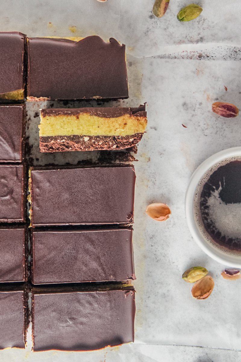 A chocolate pistachio bar sits on it's side with the three layers visible next to other bars and a cup of coffee on a gray surface.