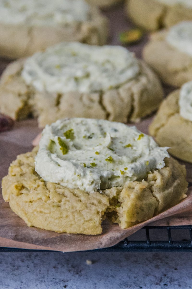 A side angle close up of a pistachio sugar cookie topped with a pistachio buttercream sits on a lined cooling rack on a light gray surface.