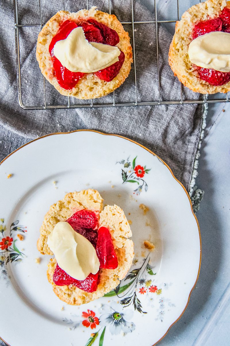 A halved scone topped with roasted strawberries and lightly whipped cream sit on a individual plate on a gray cloth and gray background with other halved and topped scones beside.
